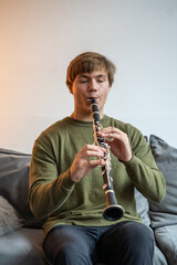 Clarinet player. Concentrated clarinetist playing flute woodwind music instrument sitting on couch at home. Musical instruments. Young man plays the oboe in living room. Relaxed mental hobby concept © DimaBerlin