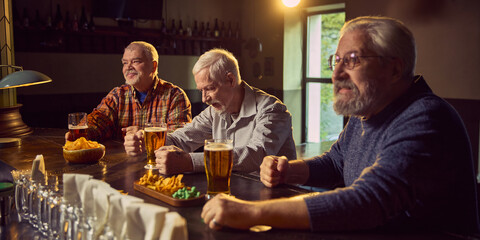 Three elderly men, friends gathering in modern pub to watching online game broadcast, drinking beer with chips, supporting favorite team. Concept of sport fans, elderly lifestyle, leisure activity