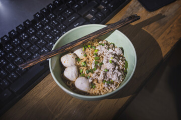 Late night Meal for working. Instant noodles flavor add nutritional value with minced pork and meatballs and topped with chopped coriander in a bowl on the desk near the keyboard.