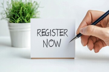 Register submission conceptual. A hand writes "REGISTER NOW" on a small card, with a potted plant in the background, emphasizing action and engagement.