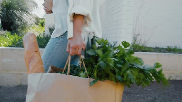 Close-up of a woman walks along a sidewalk, holding a paper bag filled with fresh herbs and a baguette in a neighborhood setting