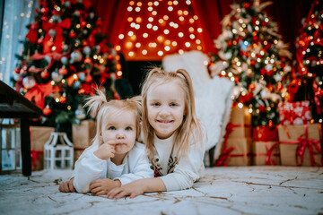 Two young siblings lying on the floor in a festive setting with Christmas trees, lights, and gifts, sharing a loving moment during the holidays..