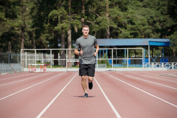 Fototapeta premium Athletic man jogging on outdoor running track in well-maintained park surrounded by tall evergreen trees, wearing casual sports attire