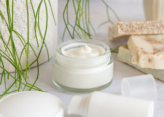 Opened cream jar with white blank lid on marble near green plant close up, cosmetic mockup