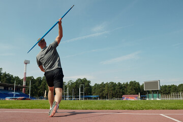 Athlete training for javelin throw on sports track with blue sky background. Sports equipment and green field visible around illustrating sports environment