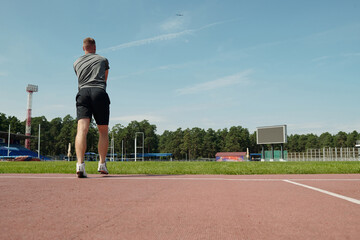 Man running on outdoor athletics track, surrounded by greenery and sports facilities. Engaging in fitness activity under clear sky without using any equipment