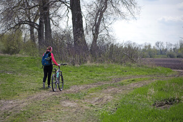 Fototapeta premium a girl with a bicycle walks a field trail. Woman riding a mountain bike in the forest. Hiker woman with backpack. one young woman riding a bike in nature. rolling the bike in his hands. back view.