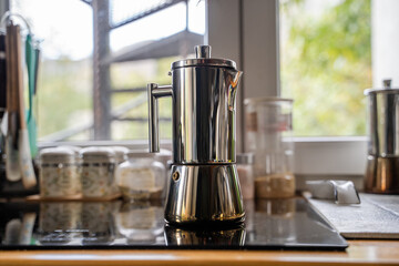 Geyser coffee maker in the home kitchen near the window. A modern stainless geyser coffee pot on an induction stove panel indoor. Coffee lovers and preparing coffee at home. Close up.