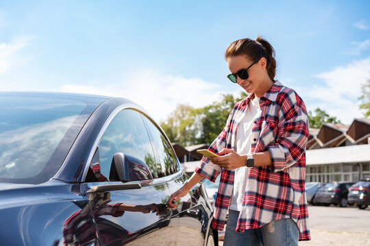 Caucasian woman in casual outfit unlocking rental car using her smart phone and mobile app outdoors in summer.