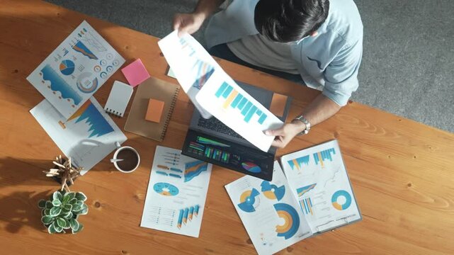 Top aerial view of manager working on laptop while looking at data analysis. Leader compare financial statistic graph while making decision at meeting table with document scatter around. Convocation.