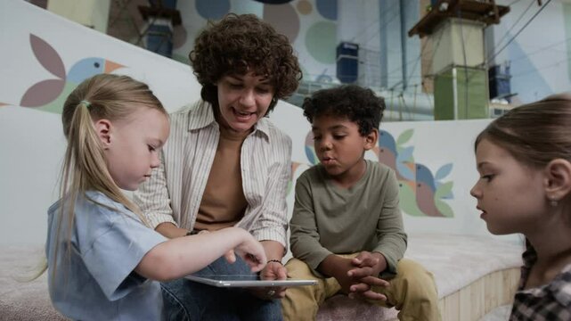 Low angle view of female Caucasian tutor drawing together with diverse kids on digital tablet in play zone