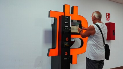 Active Senior Man Using Bitcoin ATM. The pensioner makes an automatic transaction in BTC cryptocurrency with his phone. Business, Finance, Banking, And Senior Lifestyle Using Technology Concepts