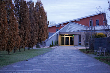 A modern building with angular architecture is surrounded by tall, brown-leafed trees and a paved...
