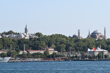 Fototapeta premium View of Topkapi Palace and Hagia Sophia from the Bosphorus, with lush greenery and historical buildings on the shore, and the Turkish flag flying near the waterfront 