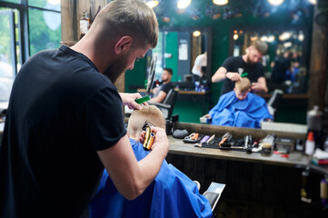 Hairdresser shaving boy's head. Competent barber using electric shaver at barbershop. Little client getting stylish men's haircut.