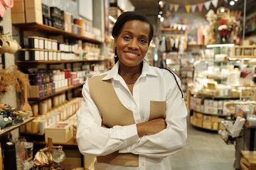 Portrait of smiling woman standing in a well-stocked store with arms crossed, surrounded by various products and decorative items in background