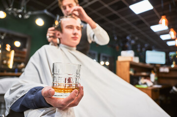 Close up photo of young client holding glass of whiskey. Male customer relaxing while getting new haircut. Professional hairdresser in fancy outfit cutting client's hair.