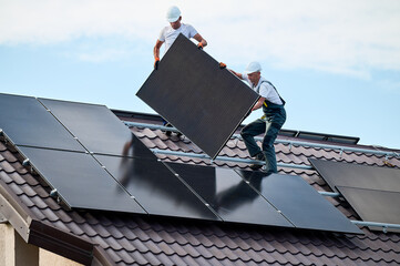 Workers building solar panel system on roof of house. Two men installers in helmets carrying...