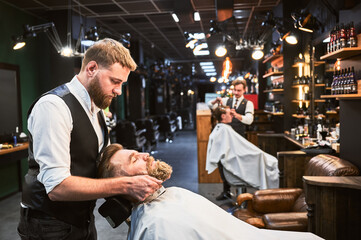 Professional barber shaving client's beard. Modern barbershop with fancy interior. Hairdresser in elegant outfit at work.