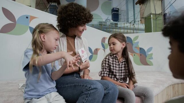 Wide shot of small kids listening to their teacher training letters with them, using paper cards on playground