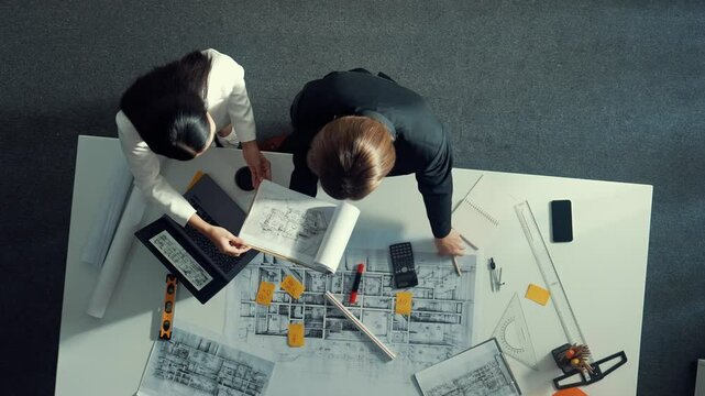 Top view of skilled project manager holding project plan and explain house design to architect engineer at meeting table with equipment, ruler, safety helmet and laptop scatter around. Alimentation.