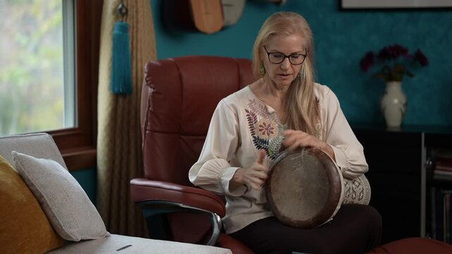 Mature woman with long blonde hair and glasses sits in a brown leather chair holding a dumbek drum looking at her phone, smiling broadly. She is wearing an ethnic blouse and black pants.