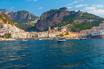 The photo captures a sunny view of Amalfi, showcasing the town's colorful buildings, steep cliffs, and vibrant coastline under a clear blue sky on a bright day.