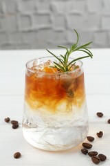 Refreshing espresso tonic drink with rosemary and coffee beans on white wooden table, closeup