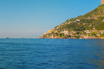 The photo captures a sunny view of Amalfi, showcasing the town's colorful buildings, steep cliffs, and vibrant coastline under a clear blue sky on a bright day.