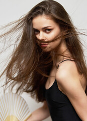 Young woman with long hair playfully holding a fan, showcasing confidence and elegance against a neutral background. Portrait and beauty concept.