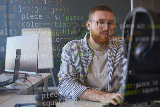 Man with glasses coding at desk in office environment, concentrating on computer screen. Reflected code illustrating work environment with technology focus