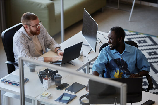 Two colleagues collaborating in a modern office, using laptops and technology to complete daily tasks. They are surrounded by desks, monitors, and office supplies