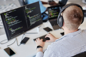 Man wearing headphones sitting at desk working on coding project on dual monitors. Image shows work...