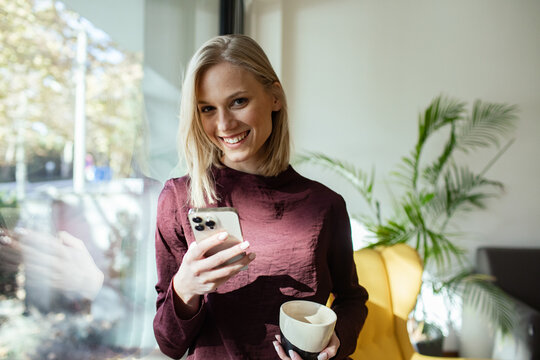 Happy woman looking at smartphone while holding coffee cup near office window
