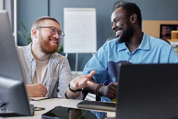 Obraz premium Two colleagues are sitting at a desk while smiling and discussing work. Laptops, documents, and other office supplies are visible in modern workspace with a bright atmosphere
