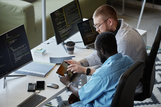 Two colleagues working together on software development in modern tech workspace, both focused on coding and using digital tools. One person is showing information on a tablet