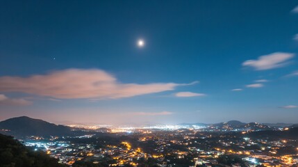 Fototapeta premium Night cityscape with bright moon and glowing city lights under blue sky. Panoramic view of illuminated urban landscape with mountains and clouds. Evening peaceful scene for tourism