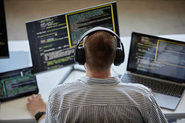 Person seen concentrating on coding projects with multiple screens and advanced technology setup wearing headset. Numerous lines of code visible across screens while typing