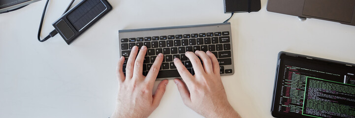 Hands typing on a wireless keyboard surrounded by various gadgets including a smartphone, power bank, and laptop suggesting tech-focused workspace