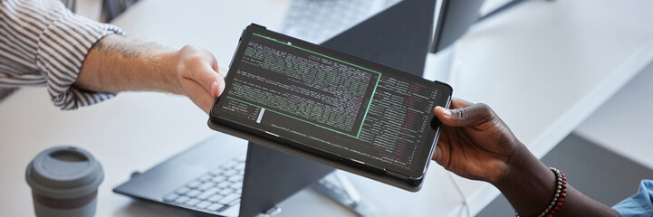Close-up of two professionals exchanging tablet displaying code in an office environment There is emphasis on collaborative effort in a tech-related project