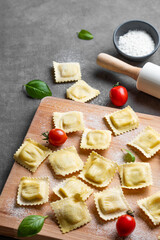 Homemade pasta ravioli with filling on wooden cutting board on dark background with tomato, flour, basil and rolling pin.
