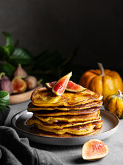 Stack of pumpkin and pancakes with honey and figs on a dark background with fresh fruits and vegetables.