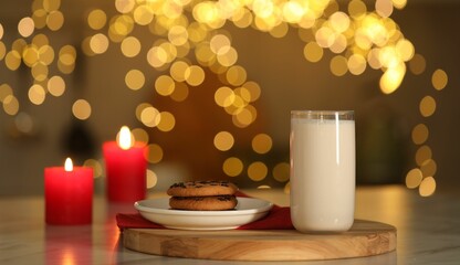 Glass of milk, tasty chocolate chip cookies and burning candles on table against blurred Christmas lights. Bokeh effect