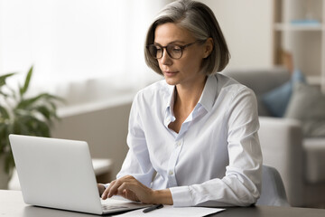 Focused pretty mature business woman in elegant glasses typing on laptop, working at computer in...