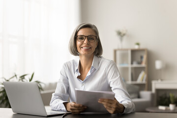 Happy successful business manager woman in glasses doing paperwork accounting job at home, working on freelance project at laptop, holding paper financial reports. Home portrait