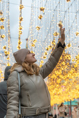 Portrait of serene adult caucasian woman in grey jacket and hat standing with raised arm on winter city street in a cold cloudy day. Glowing orange light garlands hangs between buildings.