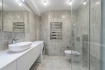 A light grey tiled bathroom with a mirrored cabinet over a white sink. Glass partition and .toilet.