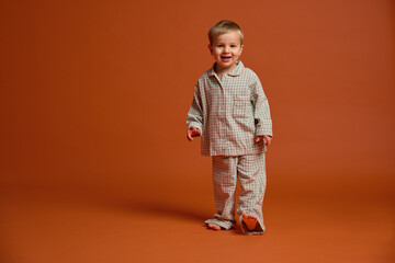 A young child smiles brightly while standing on an orange backdrop, dressed in cozy pajamas. This...