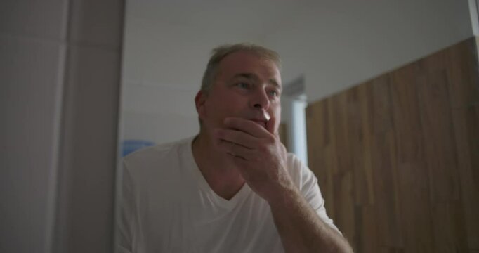 Man inspecting face in mirror, hand on cheek, checking skin after shaving, focused expression, natural wooden bathroom setting, grooming and self-care routine