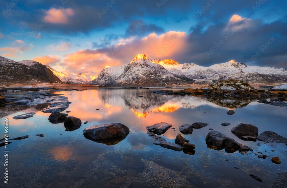 Wall mural landscape with snowy mountains reflecting on calm water with stones and colorful sky with clouds at 